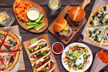 Healthy plant based fast food table scene. Overhead view on a wood background. Cauliflower crust pizzas, bean burgers, mushroom tacos, bell pepper nachos and cauliflower wings and sweet potato fries.