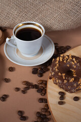 A cup of aromatic black coffee and coffee beans on the table with cinnamon sticks. Morning espresso coffee for breakfast in a beautiful white cup. With a delicious donut in the background.