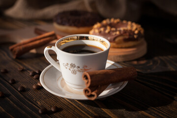 A cup of aromatic black coffee and coffee beans on the table with cinnamon sticks. Morning espresso coffee for breakfast in a beautiful white cup. With a delicious donut in the background.