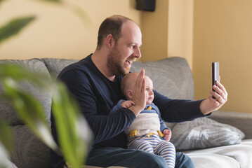 Happy father and son taking selfie with smartphone sitting on the couch at home. Portrait smiling Dad and child. Child making video call with mobile phone together at home