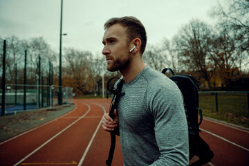 Fototapeta na wymiar Caucasian male athlete walking on track field holding backpack wearing earphones