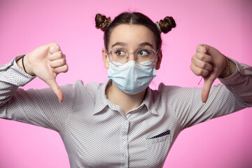Dissatisfied young teen girl in medical mask showing thumbs down unhappy with coronavirus epidemic on pink background