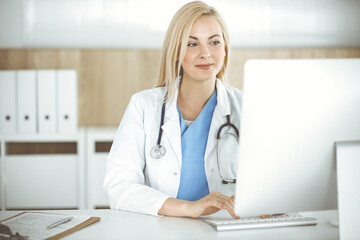 Woman-doctor at work while sitting at the desk in hospital or clinic. Blonde cheerful physician using desktop computer