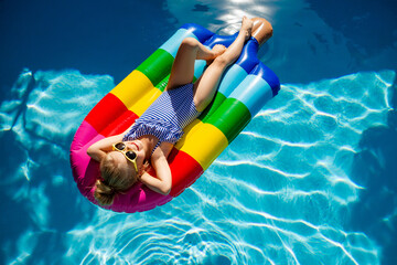 little girl in sunglasses and a striped swimsuit is lying on an inflatable multicolored striped...