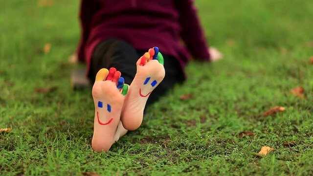 Little girl sits on grass and wounds legs which painted in funny smile faces. Summer time