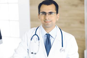 Man-doctor sitting at the desk at his working place and smiling at camera. Perfect medical service in clinic