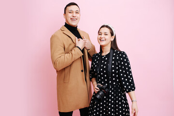 Woman photographer and a man in a coat posing and smiling on a pink background.
