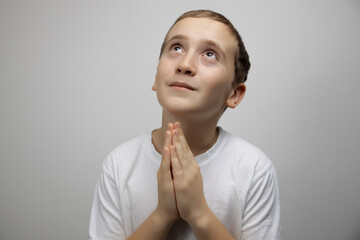 Cute child boy believer folded hands in praying gesture looking up on gray studio background, faith concept