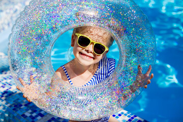 a happy little girl in sunglasses holds a swimming circle near the pool in the summer. space for text. children's summer holidays