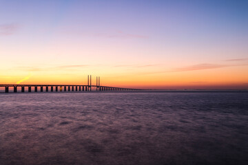 Sunset at the Oresund bridge between Sweden and Denmark at the viewpoint near Limhamn, February 2021. Wide angle, clear sky, bridge stretching from the left to the horizon, long exposure.