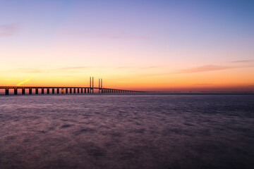Fototapeta premium Sunset at the Oresund bridge between Sweden and Denmark at the viewpoint near Limhamn, February 2021. Wide angle, clear sky, bridge stretching from the left to the horizon, long exposure.