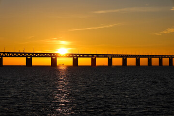 Obraz premium Sunset with the sun over and through a section of the Oresund bridge at the viewpoint near Limhamn, Sweden, in February 2021. Clear sky. Ship visible through the pillars in the evening rays.