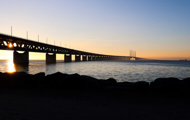 Sun seen through the Oresund bridge at the viewpoint near Limhamn, Sweden, in February 2021. Wide angle, clear sky, bridge stretching from the left to the horizon.
