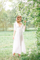 The bride in the spring in the park in a white boudoir dress. She is standing by a tree with white flowers