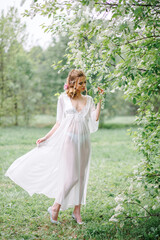 The bride in the spring in the park in a white boudoir dress. She is standing by a tree with white flowers