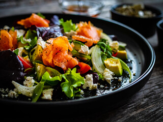 Salmon salad - smoked salmon with feta cheese, avocado and mix of vegetables on wooden table
