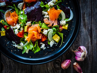 Salmon salad - smoked salmon with feta cheese, avocado and mix of vegetables on wooden table
