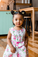 cute swarthy curly baby in a beautiful dress poses in the kitchen decorated with a garland of lamps