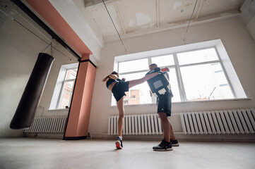Female martial arts fighter practicing leg kick or high kick with her trainer in a boxing studio at sunny day.