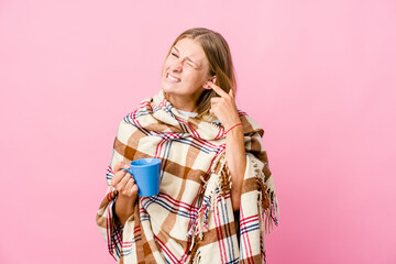 Young russian woman wrapped in a blanket drinking coffee covering ears with hands.