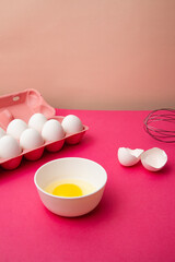 Box of eggs near bowl of yolk and whisk on pink table
