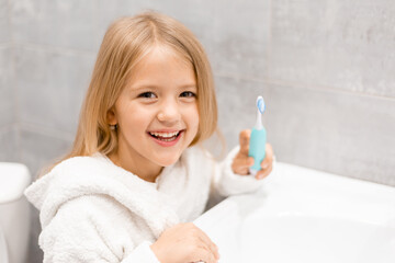 little blonde girl in a white coat holds an electric toothbrush in her hands