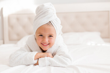 little smiling girl in a white robe with a white towel on her head is lying in bed. portrait of a child
