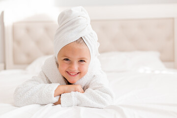 little smiling girl in a white robe with a white towel on her head is lying in bed. portrait of a child