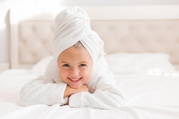 Fototapeta na wymiar little smiling girl in a white robe with a white towel on her head is lying in bed. portrait of a child