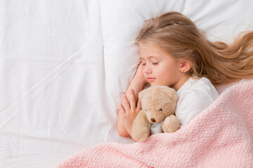 little blonde girl sleeping in bed hugging a teddy bear under a pink knitted blanket, space for text