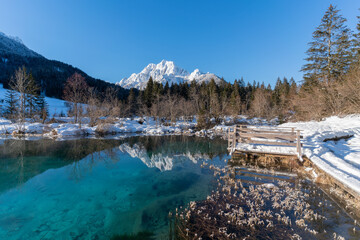 romantic path through snow along a beautiful emerald, clear volcanic lake in winter at a touristic nature reserve in the Alps. Reflection of snowcapped mountains glowing in the rising sun. no people