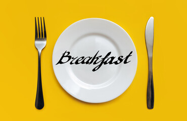 A white plate on which the word Breakfast is written standing on a yellow tablecloth . The concept of a balanced diet, ration and medical fasting. Top view, white background, copy space.