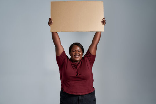 Excited plus size young african american woman in casual clothes looking at camera with a smile, holding blank cardboard banner above her head, posing isolated over gray background