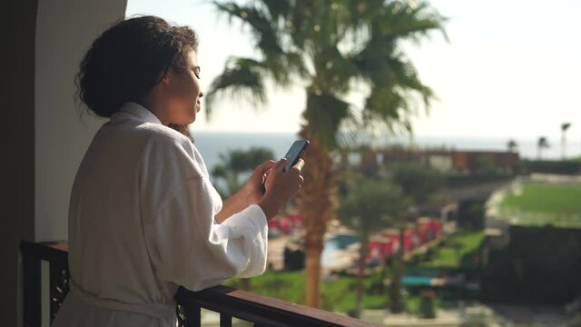 Young woman texting and liking pictures using smartphone while standing on hotel balcony outdoors with beautiful tropical landscape in the background