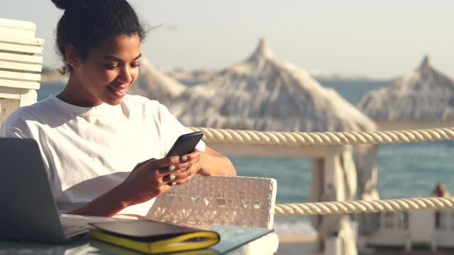 Be in touch. Joyful young woman making notes, chatting with friends and colleagues using mobile phone while relaxing on the beach resort, working remotely. Freelance work, travel concept