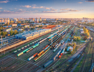 Aerial view of freight trains at sunset. Top view of railway station, wagons, railroad. Heavy industry. Industrial landscape with train in depot, city, buildings, colorful sky. Transportation 