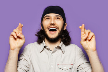 Studio portrait of excited young man, with crossed fingers and cheerful smile, on the background of purple wall.