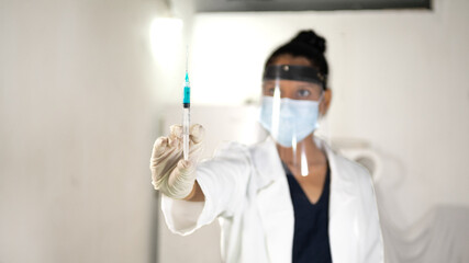 Brunette woman with protection mask, surgical gloves and medical gown in a laboratory holding a syringe with blue liquid inside that serves as a vaccine against covid and other toxic viruses