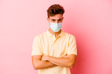 Young caucasian man wearing a protection for coronavirus isolated on pink background frowning face in displeasure, keeps arms folded.