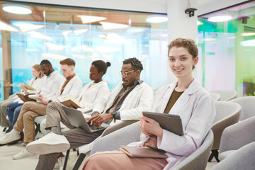Portrait of young woman wearing lab coat and smiling at camera while sitting in row with...