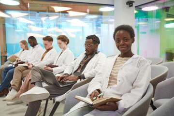 Portrait of young African American woman wearing lab coat and smiling at camera while sitting in row with multi-ethnic group of people in audience at medical seminar, copy space