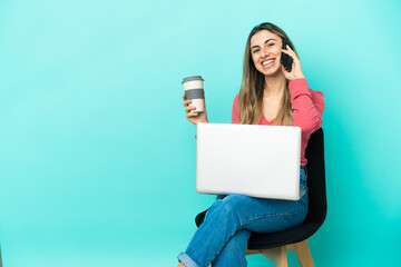 Young caucasian woman sitting on a chair with her pc isolated on blue background holding coffee to take away and a mobile