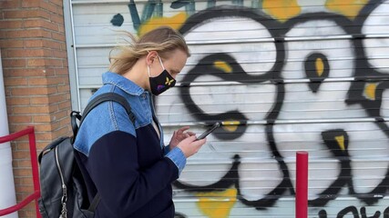 VALENCIA, SPAIN - FEBRUARY 13, 2021: Young woman in mask using mobile walking in the city against metal walls splattered with graffiti. Wearing mask became a routine, COVID-19 - Powered by Adobe