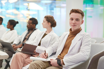 Portrait of young man wearing lab coat and smiling at camera while sitting in row with multi-ethnic...