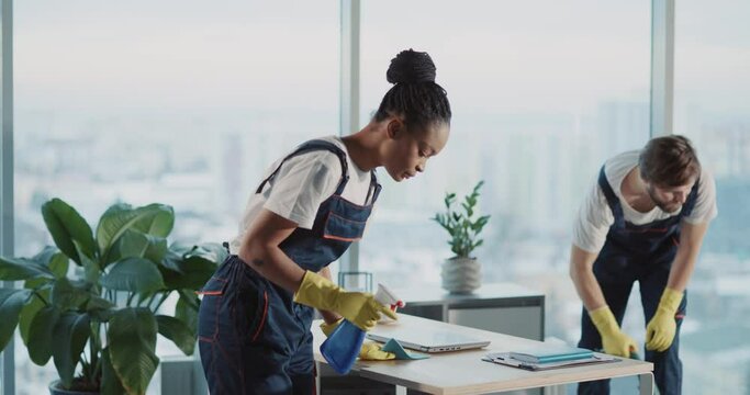Indoor portrait of afro-americna pretty young woman janitor tidying office room with colleague smiling successful at camera. Cleaning services.