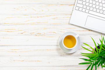 Minimalistic composition with cup of herbal tea, laptop and green flower in a pot on light wooden background. Office desktop concept. Cop space for your design. Selective focus.