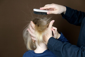 Fototapeta premium mom combing her daughter's hair, little blonde girl with a wooden brush with natural bristles, child and woman on a dark brown background, concept of hygiene, hair care, childhood, motherhood