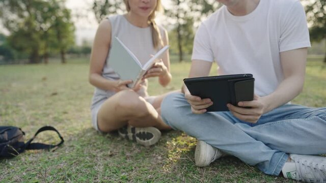 Young colleague working together on the outdoor park, sit down the ground with green grass, discuss brainstorm helping hand on the project, using tablet doing research, classmate reviewing books 