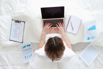Woman sitting in bed with documents and laptop top view