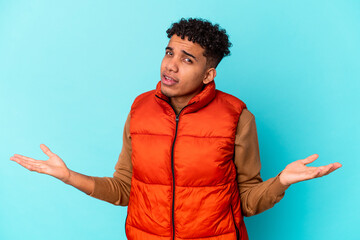 Young african american curly man isolated on blue doubting and shrugging shoulders in questioning...